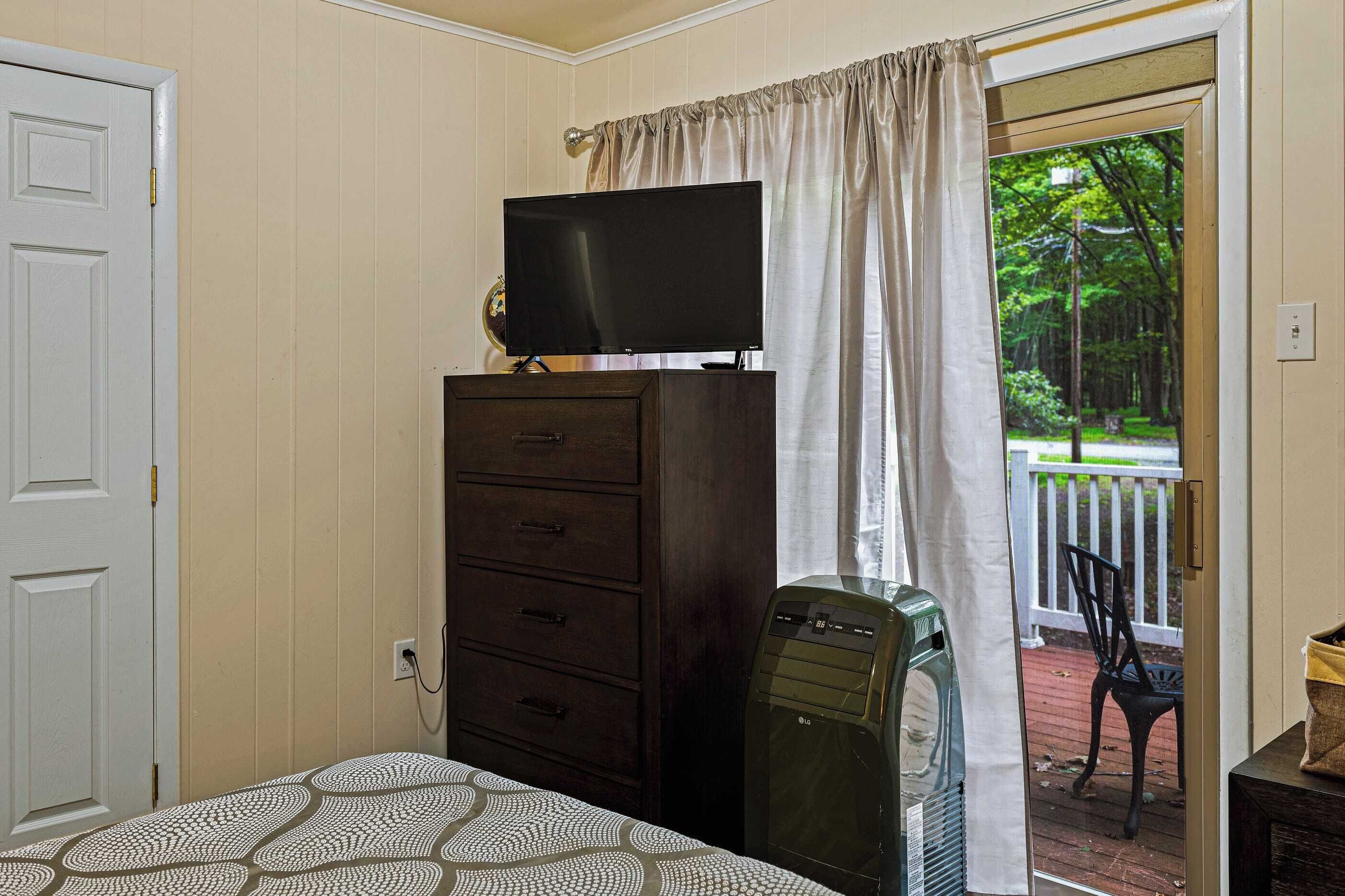 A bedroom with a bed, a dresser, a television on top, a heater, and a view of a deck with a chair.