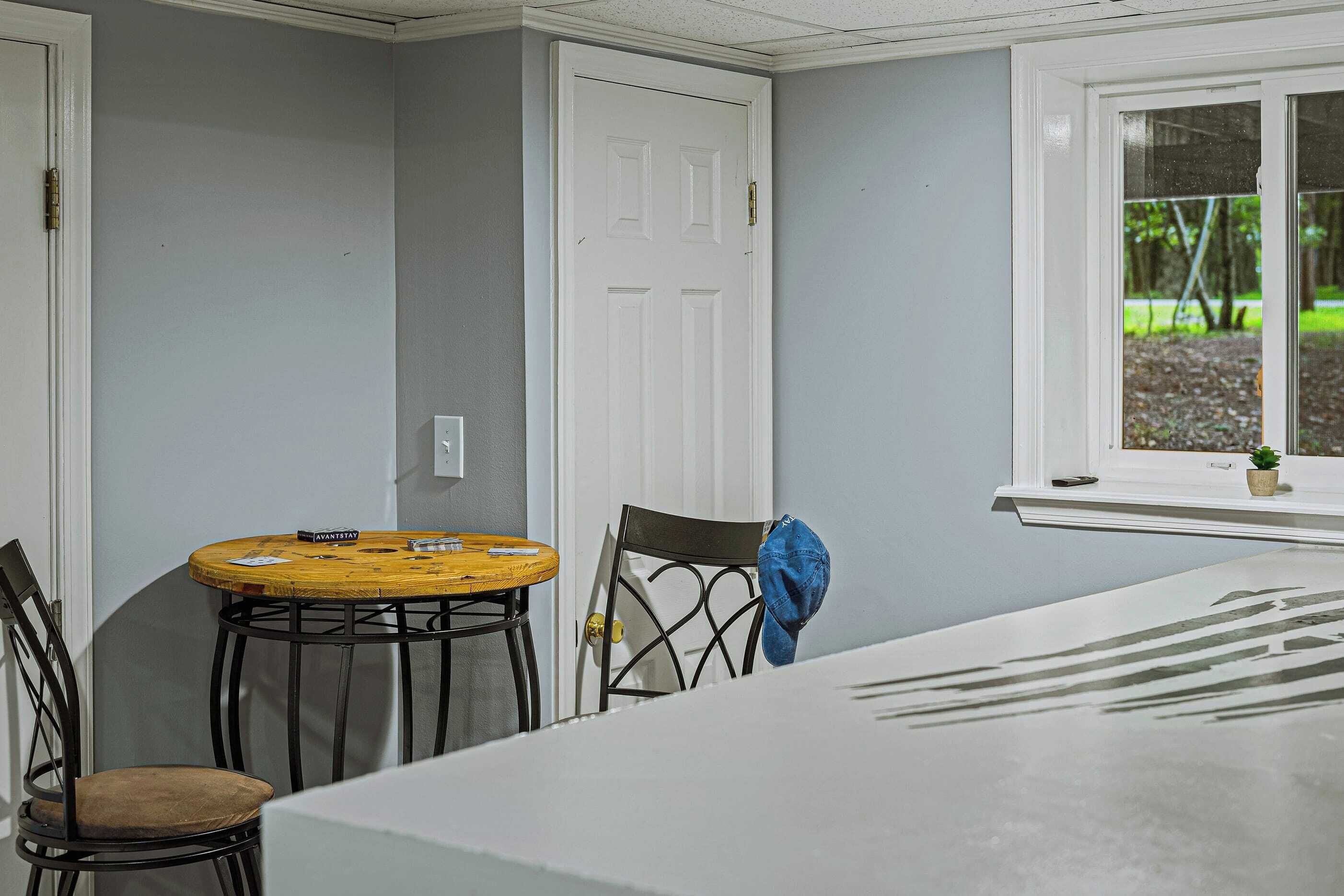Ein Zimmer mit einem weißen Tisch, zwei Stühlen, einem Holzstuhl, einer Tür und einem Fenster mit Blick auf Grünflächen.
