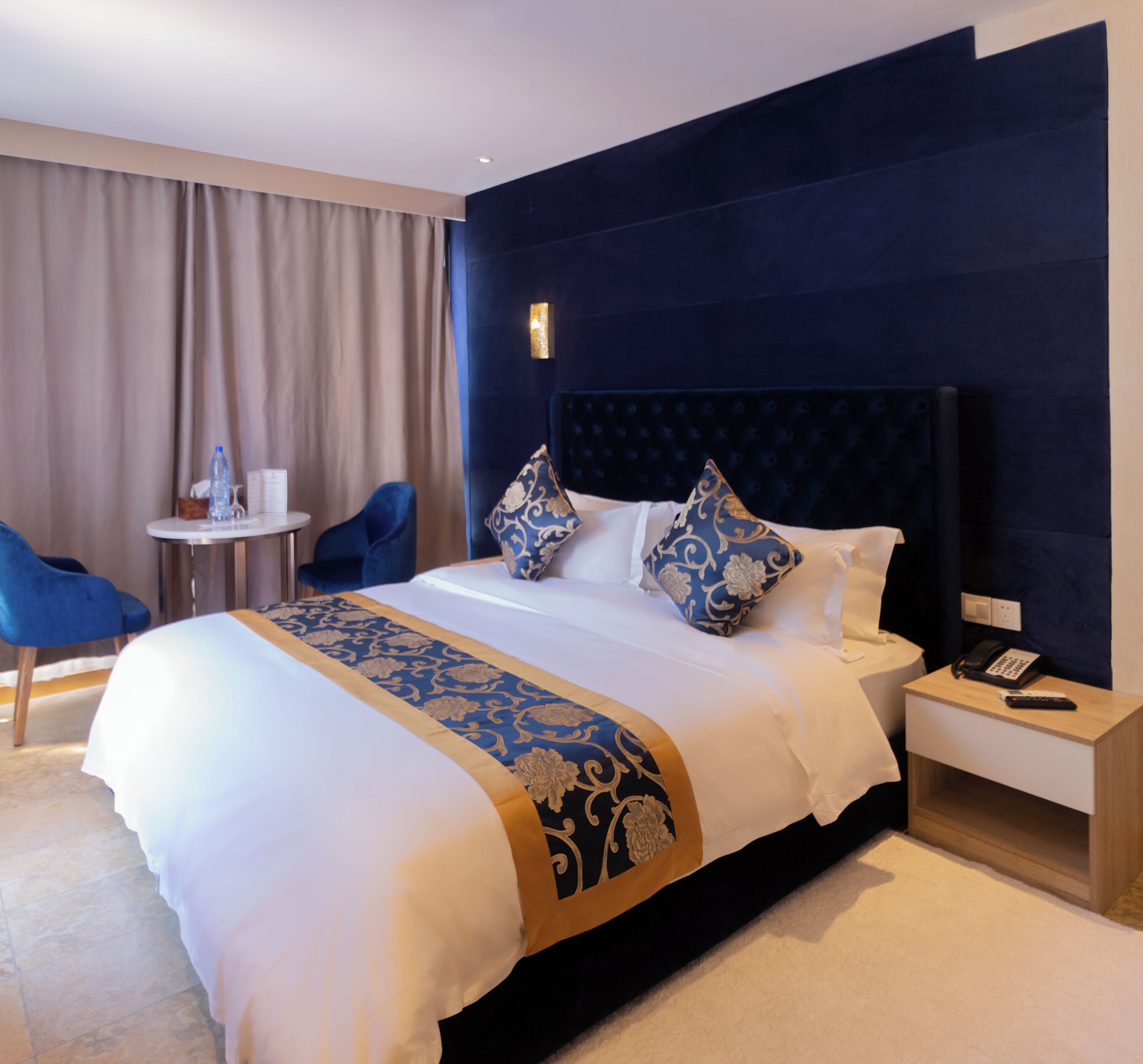 Ein Hotelzimmer mit einem großen Bett, einem Nachttisch mit Telefon, einem Sessel und einem kleinen Tisch mit Wasserflasche.