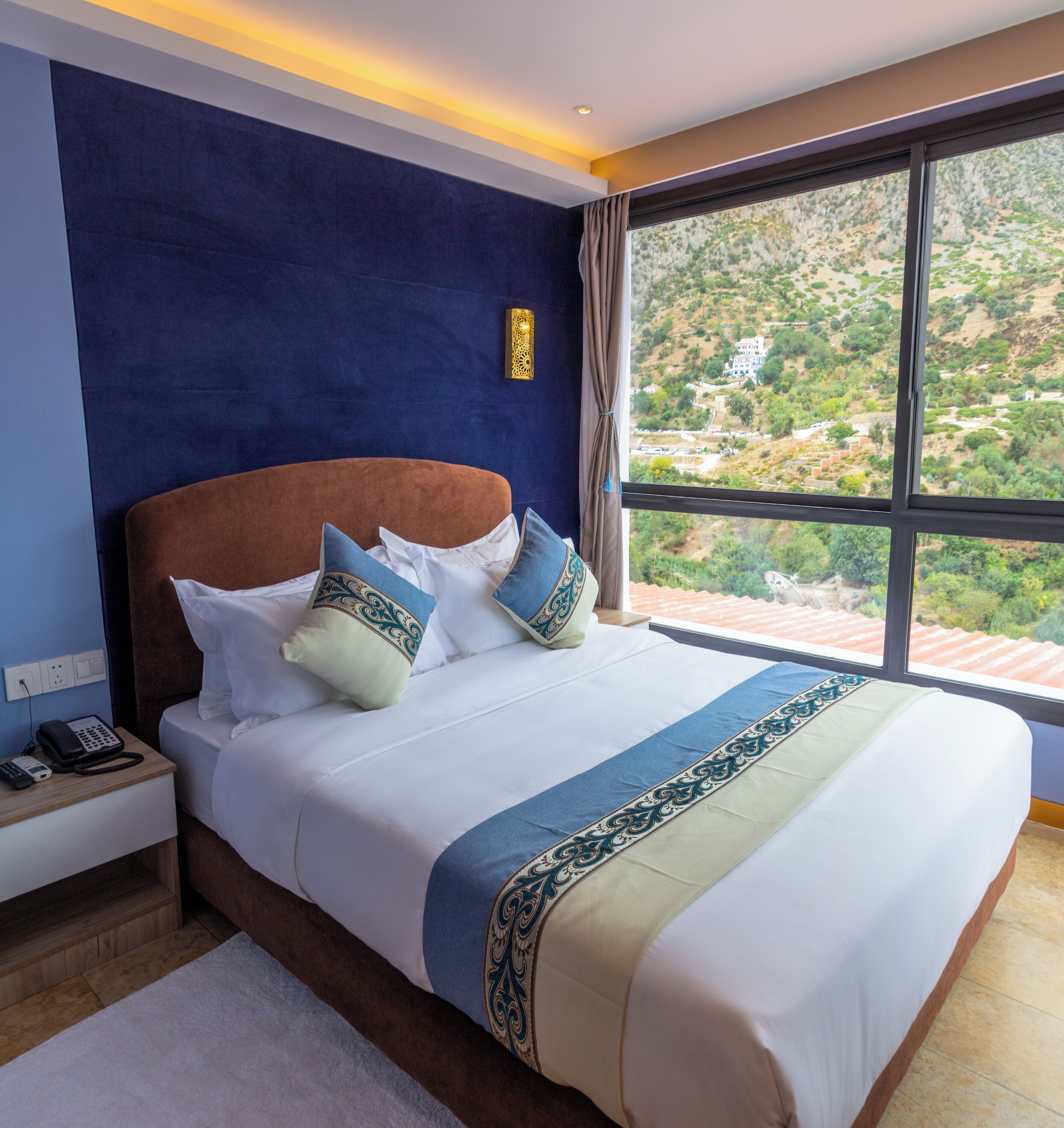 Ein Hotelzimmer mit einem großen Bett, einem Nachttisch mit Telefon und Blick auf eine hügelige Landschaft durch ein großes Fenster.