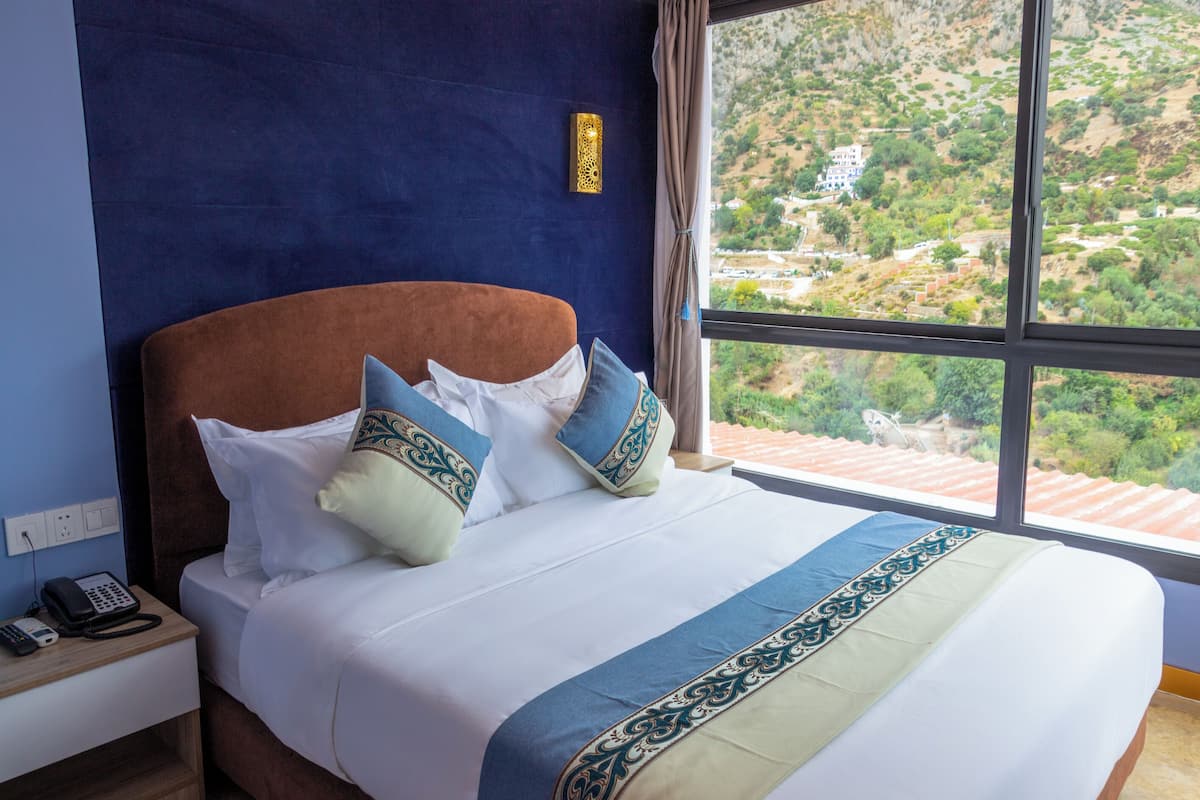 A hotel room with a large bed, a nightstand with a phone, and a view of a hilly landscape through a large window.