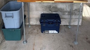 Eine Betonmauer mit einem Metallregal, einem Holztisch, einem blauen Kühlbox und einer Mülltonne.