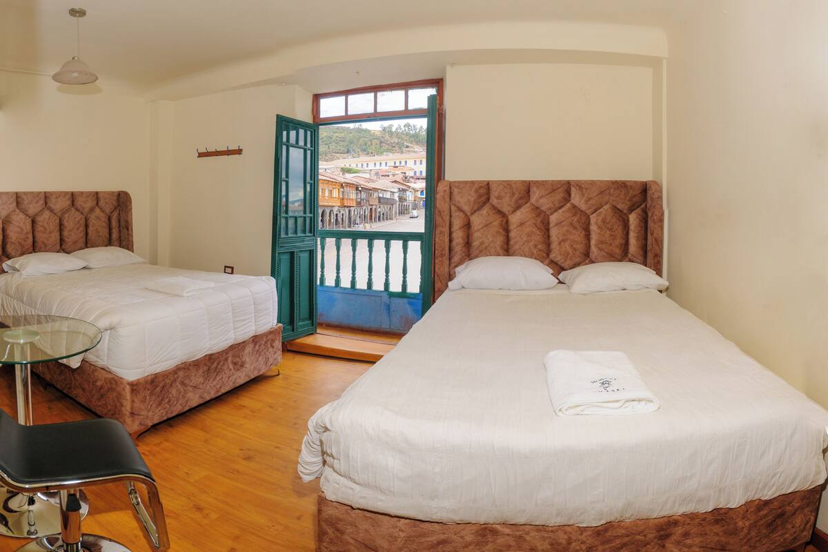 A room with two beds, a glass table, a chair, and a view of a street through an open door.