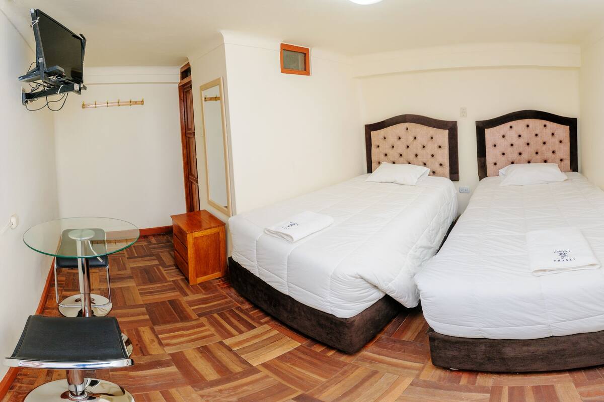A hotel room with two beds, a glass table, a wooden floor, and a TV mounted on the wall.