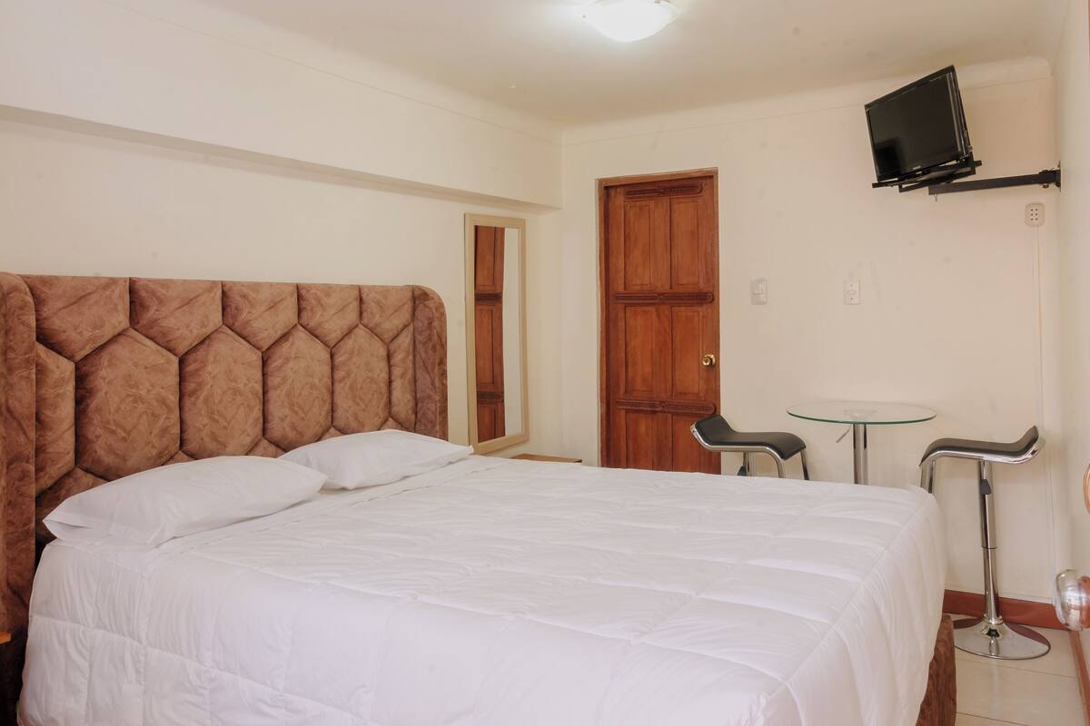 A bedroom with a large bed, a wooden door, a small table, and a TV mounted on the wall.