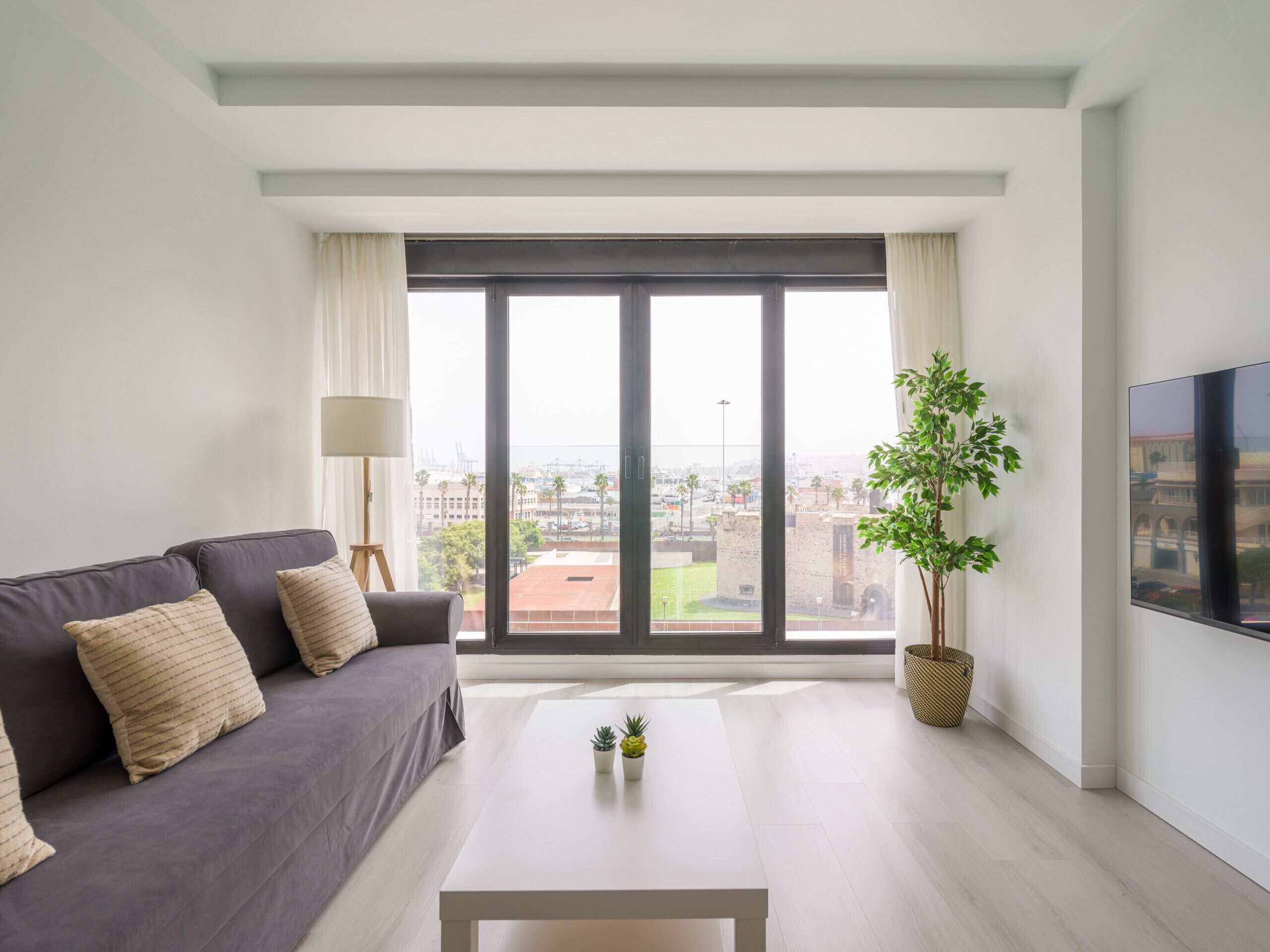 Ein modernes Wohnzimmer mit einem grauen Sofa, einem weißen Couchtisch und einem großen Fenster mit Blick auf die Stadt.