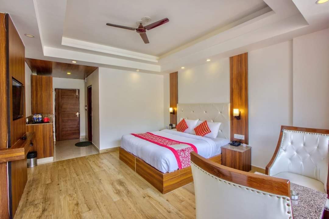 A modern hotel room with a wooden floor, a large bed with white and red bedding, a white leather armchair, a wooden side table, and a ceiling fan.