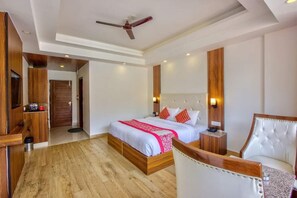 A modern hotel room with a wooden floor, a large bed with white and red bedding, a white leather armchair, a wooden side table, and a ceiling fan. - Hotel The Ventus - A boutique Hotel  (New Delhi)