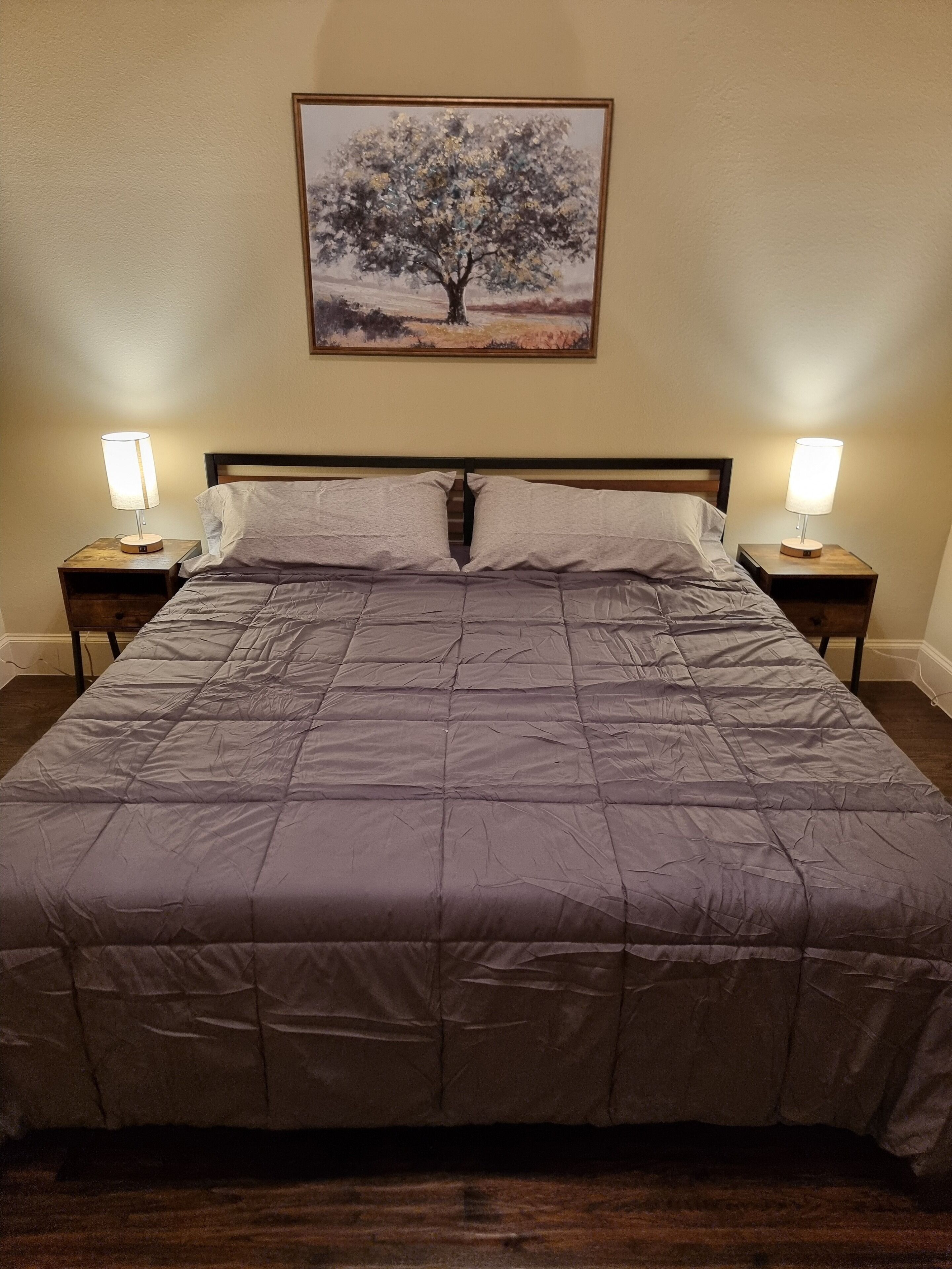 A bed with a gray quilt, two bedside tables with lamps, and a painting of a tree on the wall.