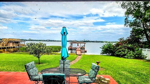 Un patio con una mesa redonda y sillas, una sombrilla azul, y vistas a un lago con un muelle y un pabellón.