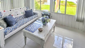 Habitación luminosa con sofá blanco, una mesita de centro y grandes ventanales con vistas a una zona verde y un edificio.