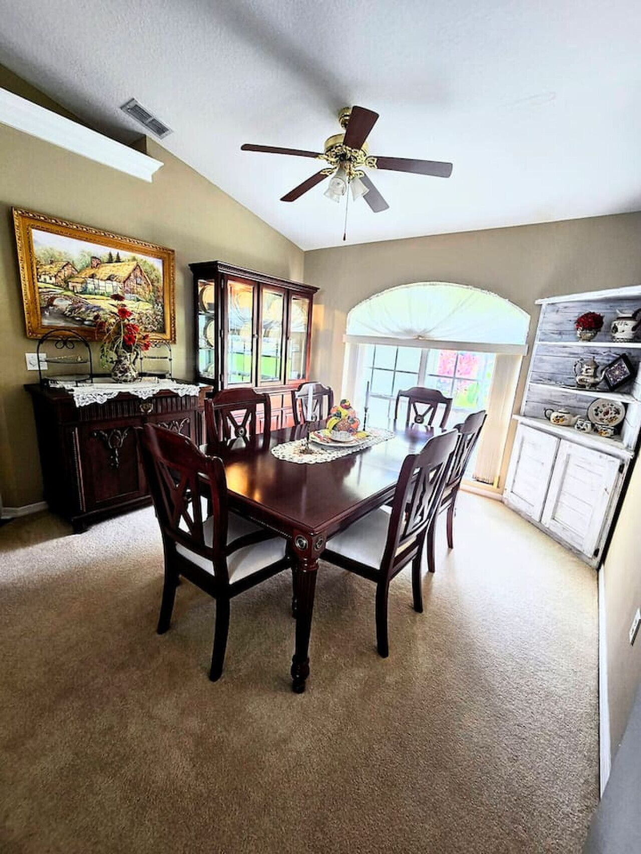 A dining room with a wooden table and chairs, a ceiling fan, a cabinet with glass doors, and a painting on the wall.
