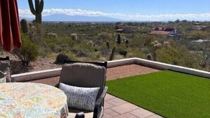 A patio with a table and chairs, a cactus, and a view of a desert landscape with mountains in the distance.