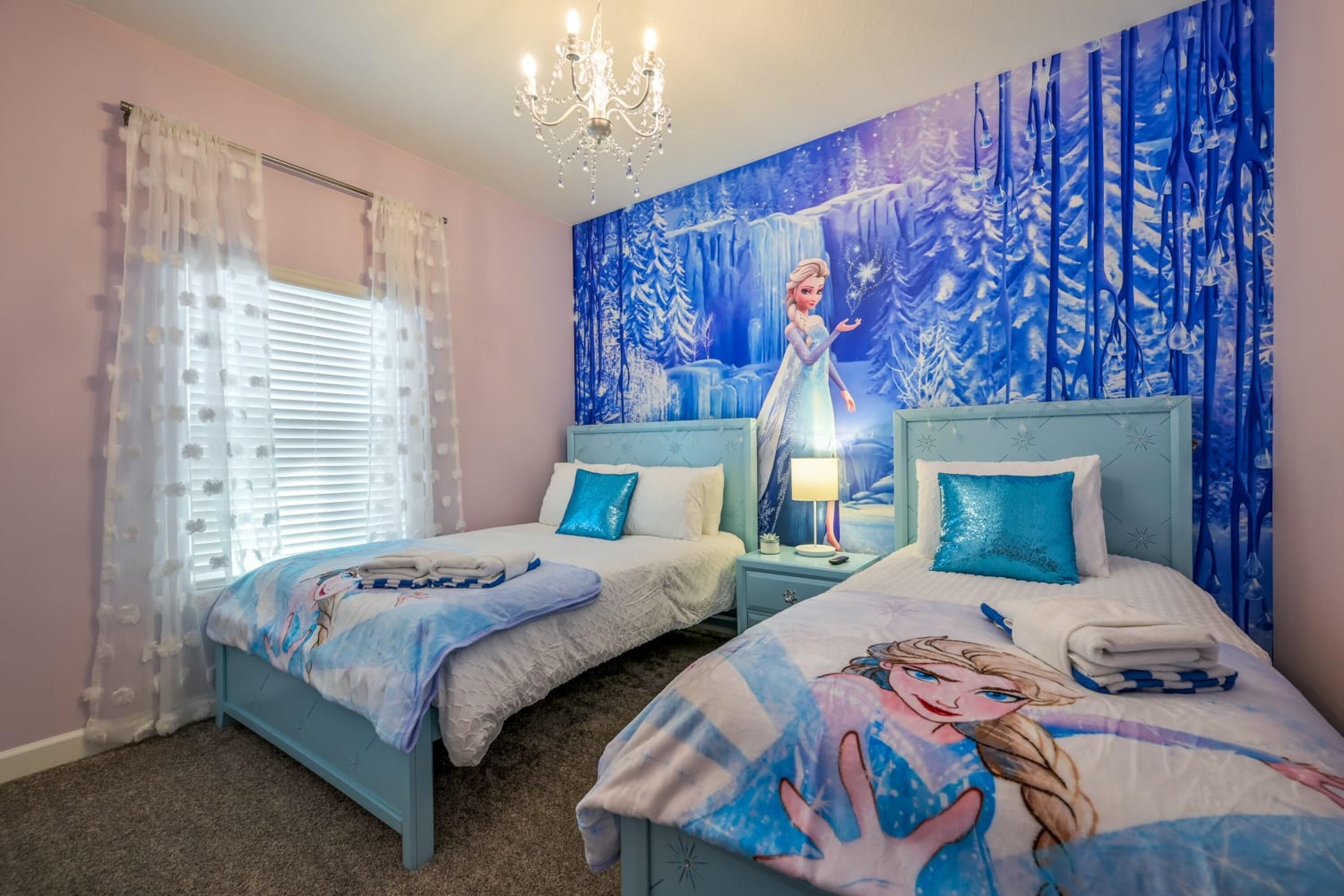 A bedroom with two beds, each with a blue headboard and a Frozen-themed bedspread. There is a large mural on the wall depicting a winter scene with a character.