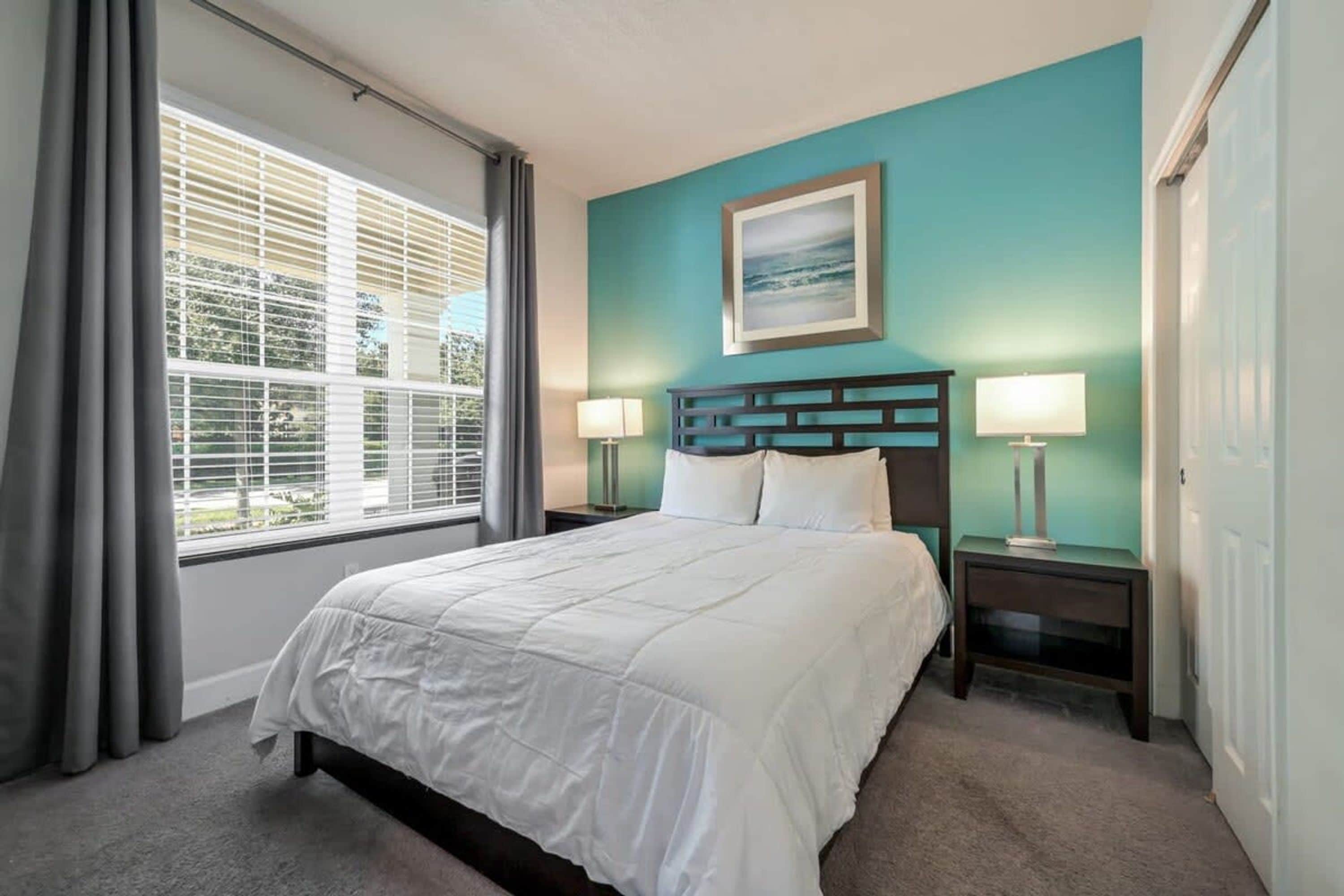A hotel room with a large bed, two bedside tables with lamps, a window with blinds, and a teal-colored wall.