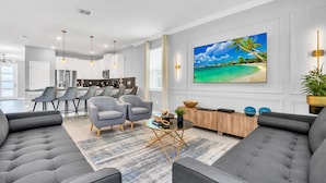 A modern living room with a large flat-screen TV, grey sofas, a coffee table, and a dining area with bar stools. - Luxury Incredible Themed Home Best Vacation (Kissimmee)