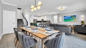 A modern kitchen with a central island, a dining area with a wooden table set for six, and a living room with a gray sofa and a large TV. - Luxury Incredible Themed Home Best Vacation (Kissimmee)