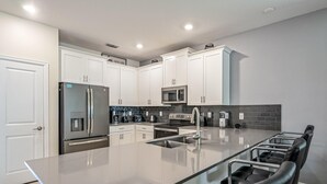 A modern kitchen with white cabinets, stainless steel appliances, a large island with seating, and a tiled backsplash. - Family Paradise Space Themed Perfect Location (Kissimmee)