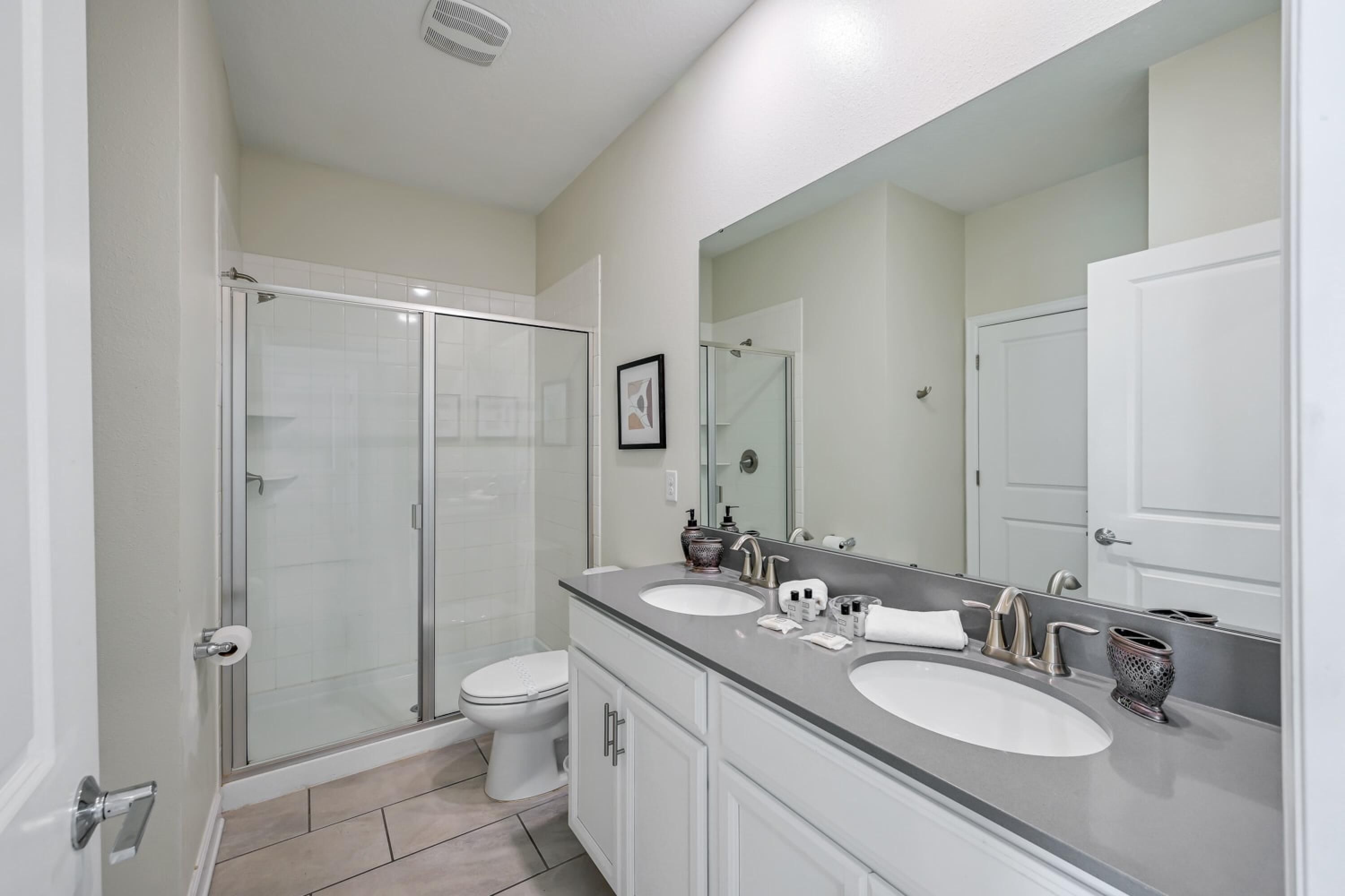 A modern bathroom with a glass shower enclosure, a white toilet, and a vanity with two sinks and a large mirror.