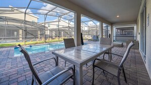 A covered patio with a glass table and chairs, a pool, and a view of a house through a greenhouse structure. - Family Paradise Space Themed Perfect Location (Kissimmee)