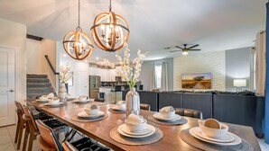 A modern dining area with a long wooden table set for six, surrounded by leather chairs. There are two pendant lights above the table, and a kitchen is visible in the background. - Family Paradise Space Themed Perfect Location (Kissimmee)