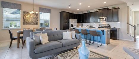 A modern kitchen with dark cabinets, a central island, and a dining area with a gray sofa and chairs.