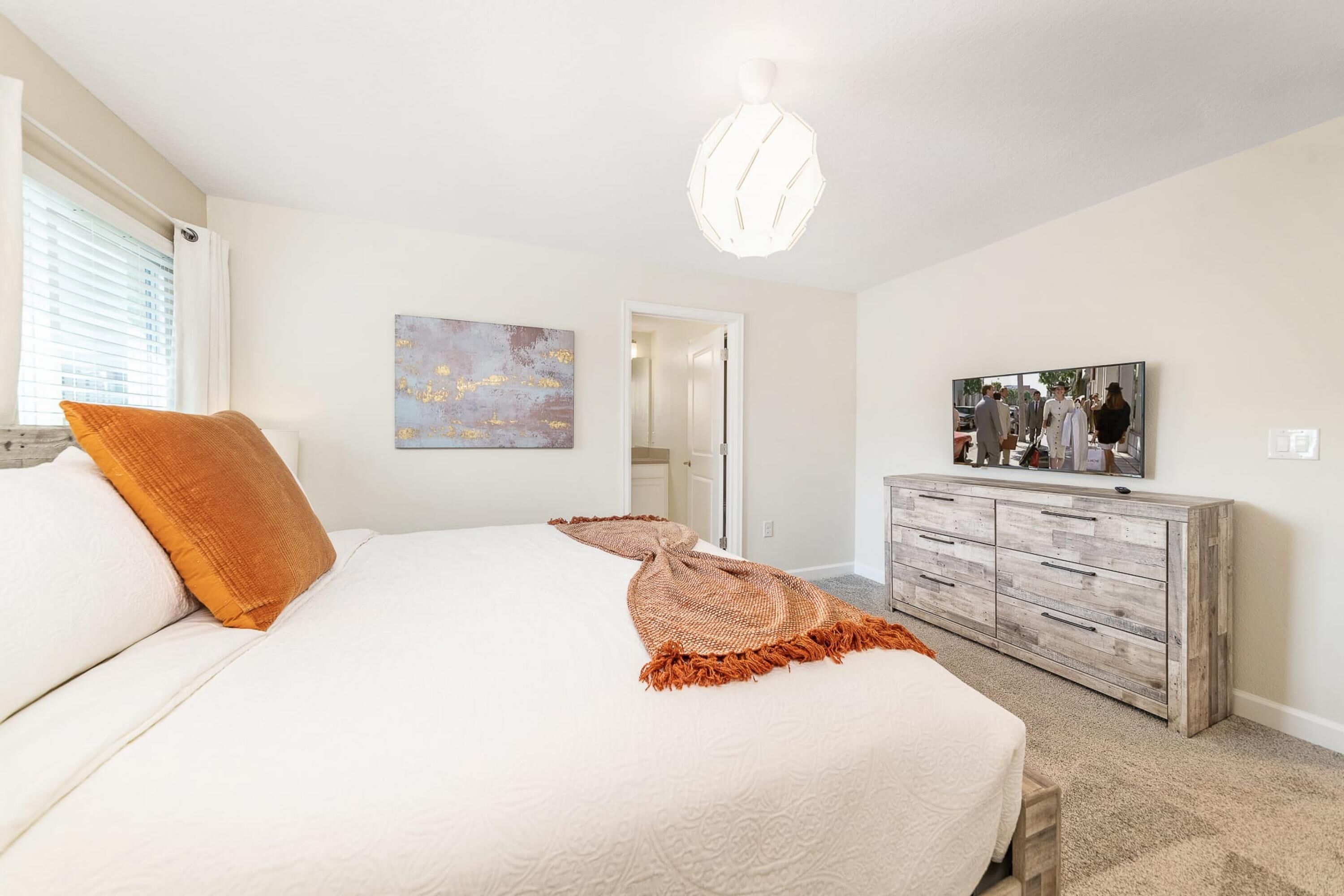 A bedroom with a large bed, a wooden dresser, a painting on the wall, and a ceiling light fixture.