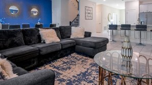 A modern living room with a black sectional sofa, a glass coffee table, and a blue patterned rug.