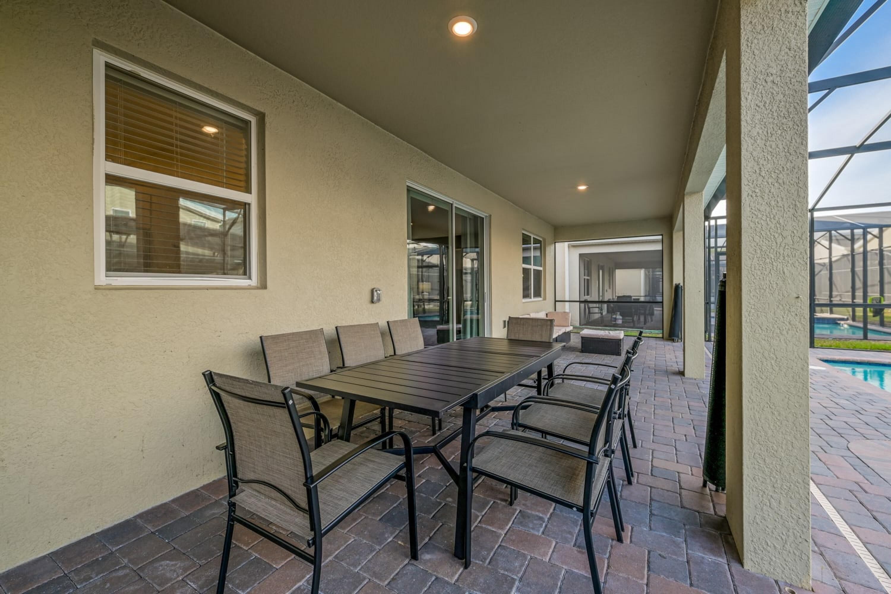 A covered patio with a long dining table and chairs, large windows, and a view of a swimming pool.