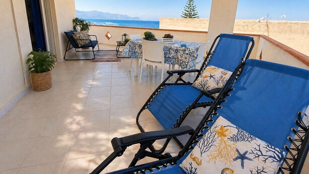 Un balcón con vista al mar, una silla de descanso, una mesa con mantel estampado y una planta en maceta.