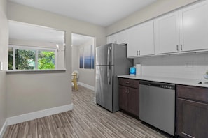 A modern kitchen with stainless steel appliances, white cabinetry, and a view through an open door to a room with a window overlooking greenery. - Stunning Stay in Rochester Hills (Rochester Hills)