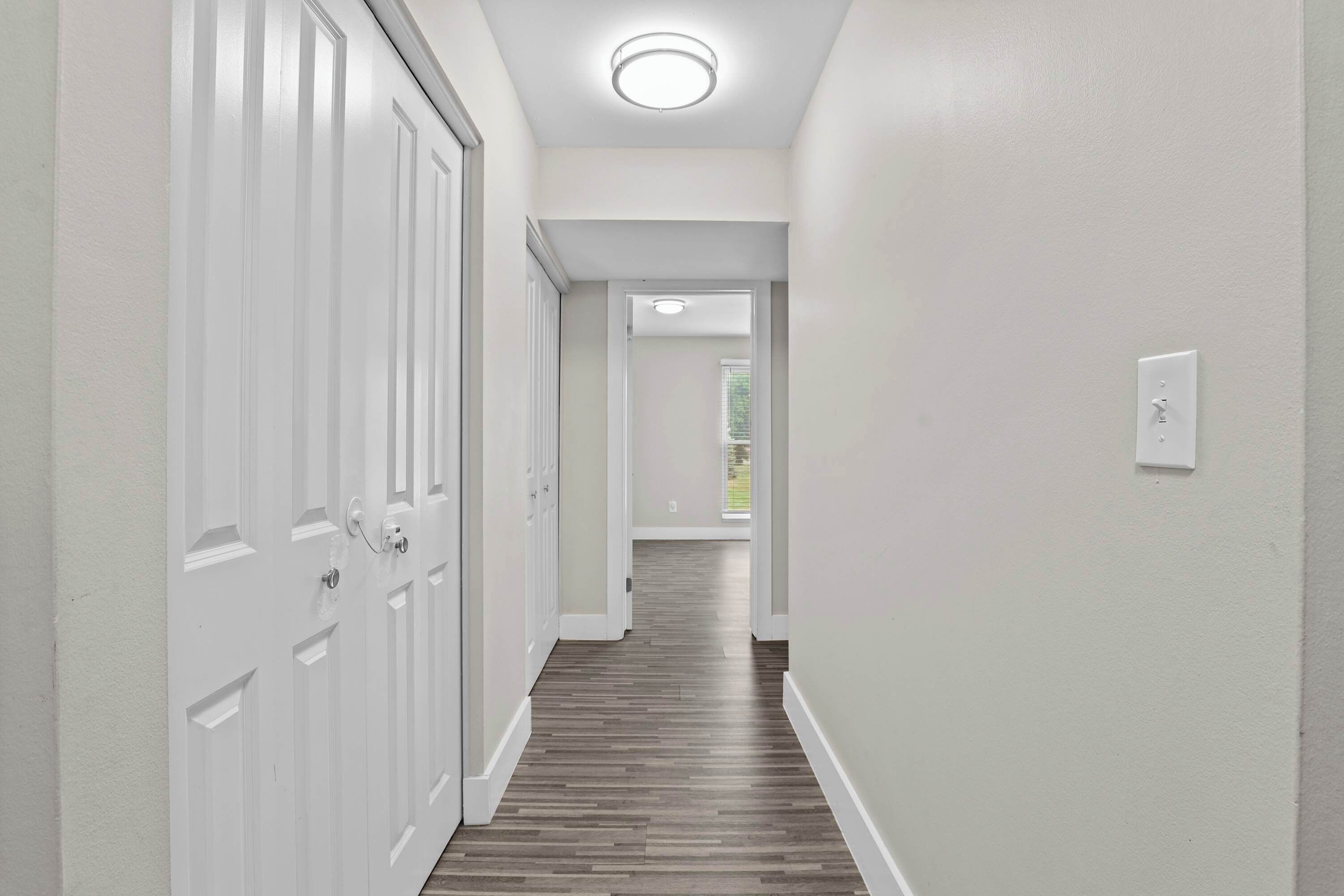 A hallway with white walls and wooden flooring leading to a door with a window.