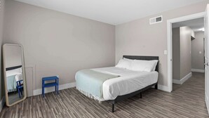 Un dormitorio con una cama, una pequeña silla azul, un espejo y una puerta que da a otra habitación.