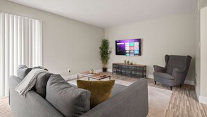 A modern living room with a grey sofa, a coffee table, a grey armchair, a TV mounted on the wall, and a potted plant. - Modern Comfort in Rochester, Perfect Getaway (Rochester Hills)