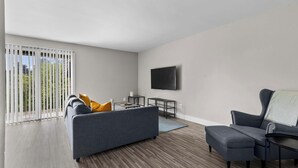 A modern living room with a grey sofa, a grey armchair, a flat-screen TV mounted on the wall, and a wooden floor. - Getaway in the Heart of the City! (Rochester Hills)