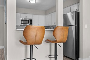 A kitchen with white cabinets, a stainless steel refrigerator, and two brown leather bar stools. - Getaway in the Heart of the City! (Rochester Hills)