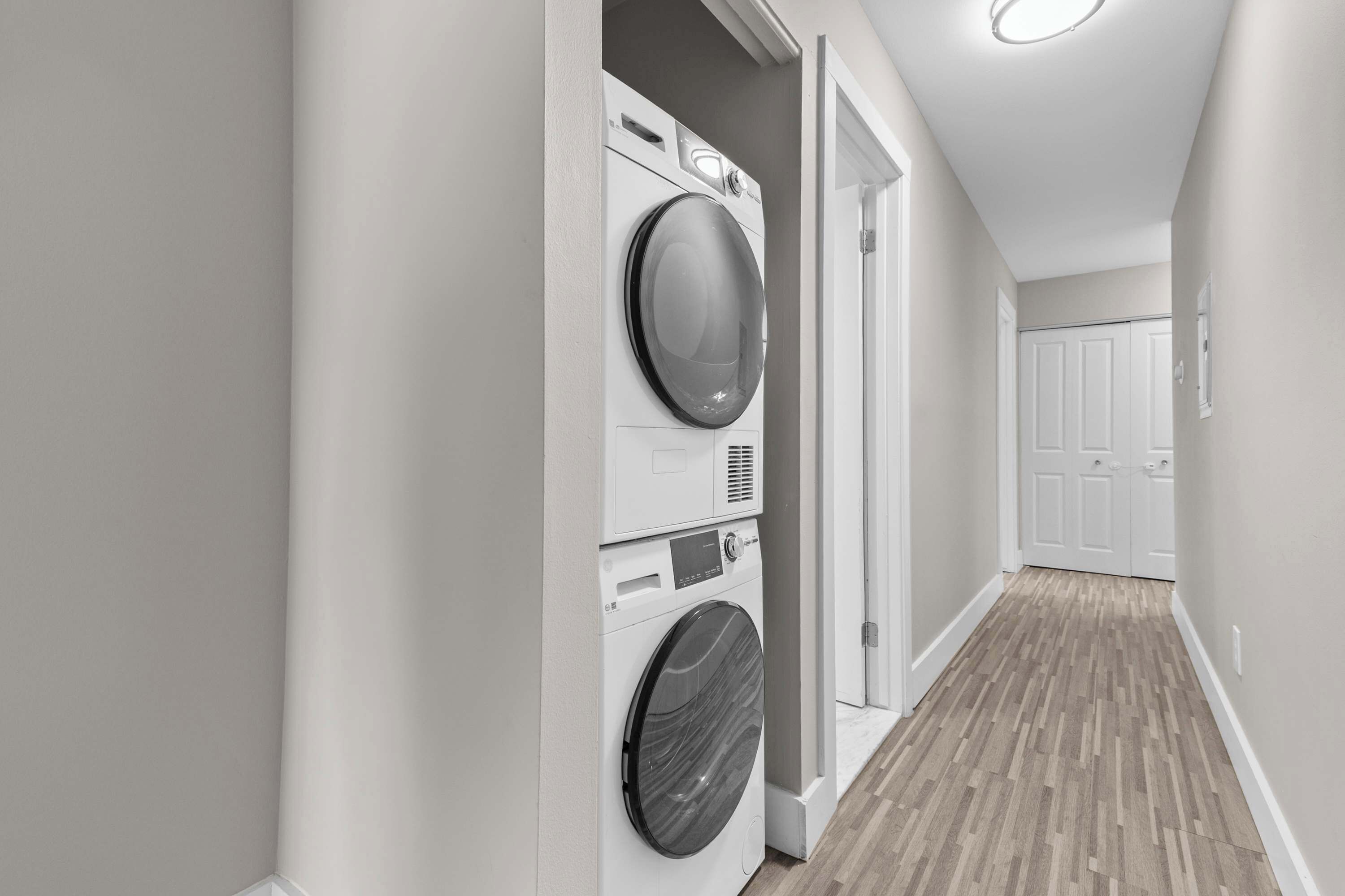 A laundry room with stacked washers and dryers, a hallway with wooden flooring, and a door leading to another room.