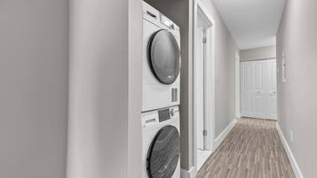 A laundry room with stacked washers and dryers, a hallway with wooden flooring, and a door leading to another room.