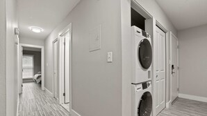 A laundry room with a front-loading washer and dryer, a hallway with a striped floor, and a door leading to a bedroom. - Spacious Stay In with Great Amenities! (Rochester Hills)