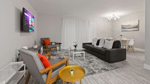 A modern living room with a flat-screen TV, a grey sofa, a wooden chair, a small coffee table, and a dining area with a white table and chairs. - Trendy & Vibrant Unit City Experience (Rochester Hills)