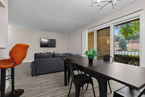 A modern living room with a grey sofa, a dining table with chairs, a flat-screen TV mounted on the wall, and a large window with a view of the outside. - The Best Stay with Parking and Amenities! (Rochester Hills)