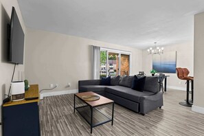 A modern living room with a sectional sofa, a coffee table, a flat-screen TV, and a large window with a view of the outside. - The Best Stay with Parking and Amenities! (Rochester Hills)