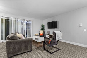 Un salon moderne avec un canapé gris, une petite table basse blanche, un fauteuil de bureau noir, un téléviseur à écran plat fixé au mur et une porte-fenêtre coulissante avec des stores verticaux.