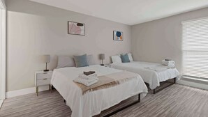A modern bedroom with two beds, a nightstand with a lamp, and a window with blinds. - Modern 2BR Retreat w/Pool & Gym | Near Downtown (Rochester Hills)