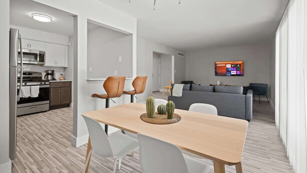 A modern kitchen with white cabinets, a built-in oven, and a microwave. There is a dining area with a wooden table and white chairs. A living room with a gray sofa and a TV mounted on the wall is visible in the background. - Peaceful Rochester Escape Perfect for Families (Rochester Hills)