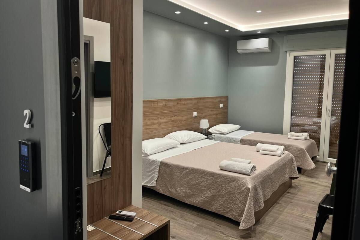A modern hotel room with two beds, a wooden headboard, a small bedside table, a wall-mounted TV, and an air conditioning unit.