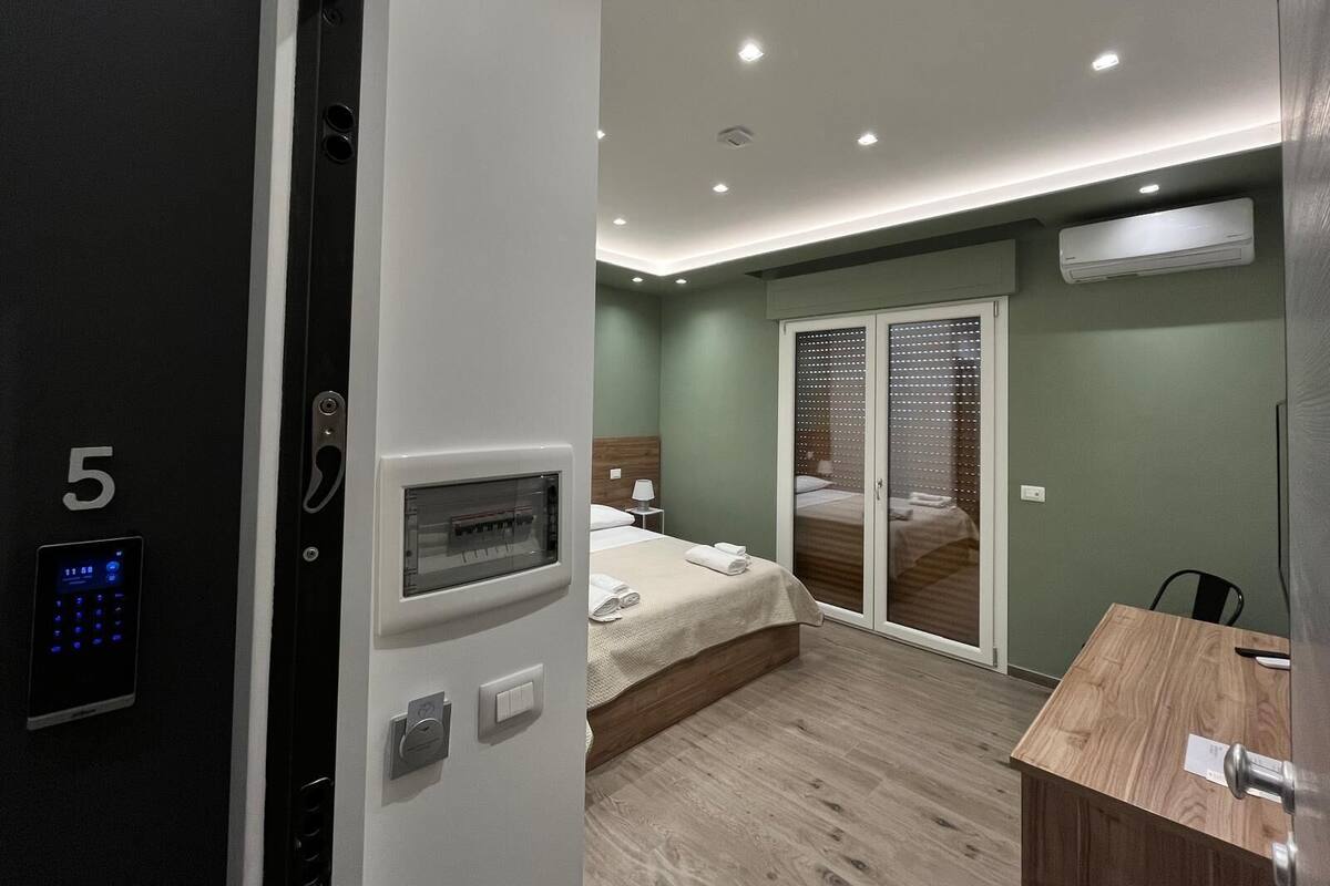 A modern hotel room with a digital keycard reader, a bed with a wooden headboard, a small bedside table, and a sliding glass door leading to a balcony.