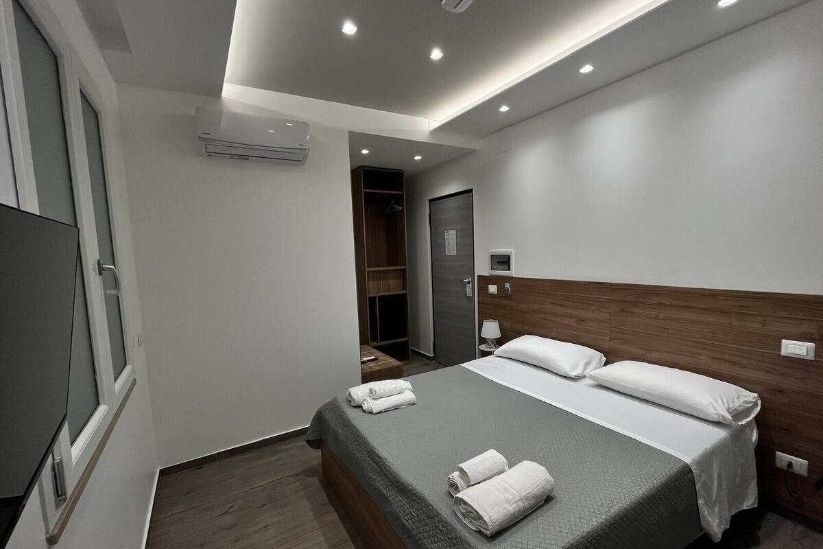A modern hotel room with a large bed, a wardrobe, a desk, and an air conditioning unit.