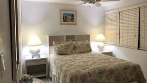 A bedroom with a bed, two bedside lamps, a ceiling fan, a nightstand, and a window with blinds.