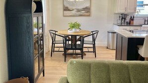 A living room with a green sofa, a dining area with a table and chairs, a kitchen with white cabinets, and a large window.